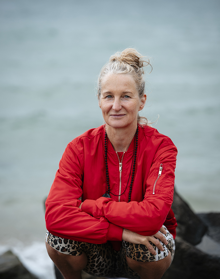 fotographie by florence - portrait of a yoga teacher wearing a red jacket, sitting on a rock at the Oriental Bay beach  in a relaxed manner looking directly at the camera with a small smile on her face
