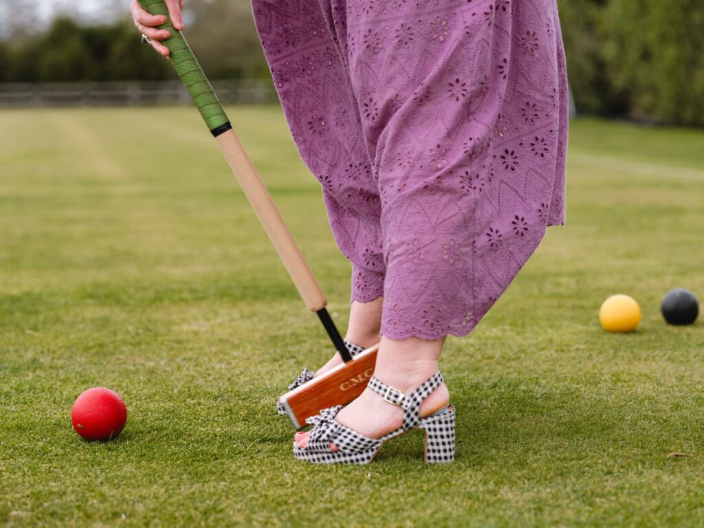 Photo by Florence Charvin Photography of Person in purple eyelet trousers and chequered heels prepares to hit a red croquet ball with a mallet on a grassy lawn, with other coloured balls nearby.