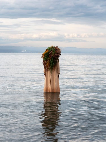 Photo by Florence Charvin Photography of A person in a long, light-coloured garment stands knee-deep in water, carrying a large bundle of seaweed on their back under a cloudy sky.