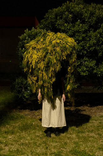 Photo by Florence Charvin Photography of A person stands outdoors at night with their upper body covered by a large bundle of leafy, yellow-flowered branches, obscuring their face and torso.