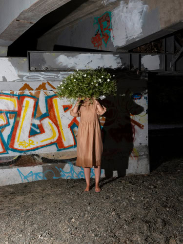 Photo by Florence Charvin Photography of A person in a peach dress stands barefoot under a bridge, holding a large bouquet of green foliage and white flowers that obscures their face; graffiti covers the wall behind them.