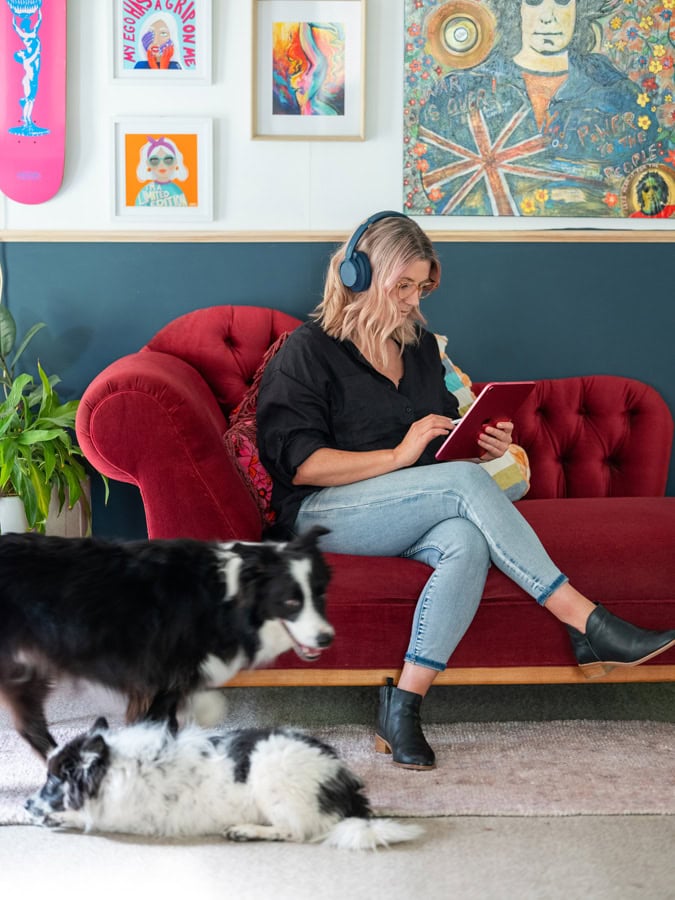 Photo by Florence Charvin Photography of A woman with headphones sits on a red sofa using a tablet, whilst two black and white dogs are nearby. Colourful artwork is displayed on the wall behind her.