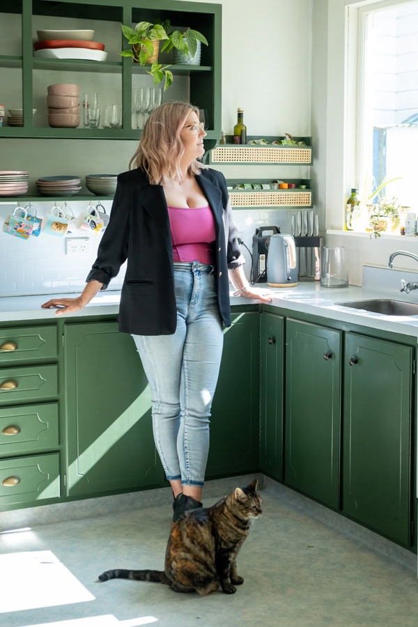 Photo by Florence Charvin Photography of A woman in a black blazer and jeans stands in a green kitchen by a window, with a cat sitting on the floor near her feet.