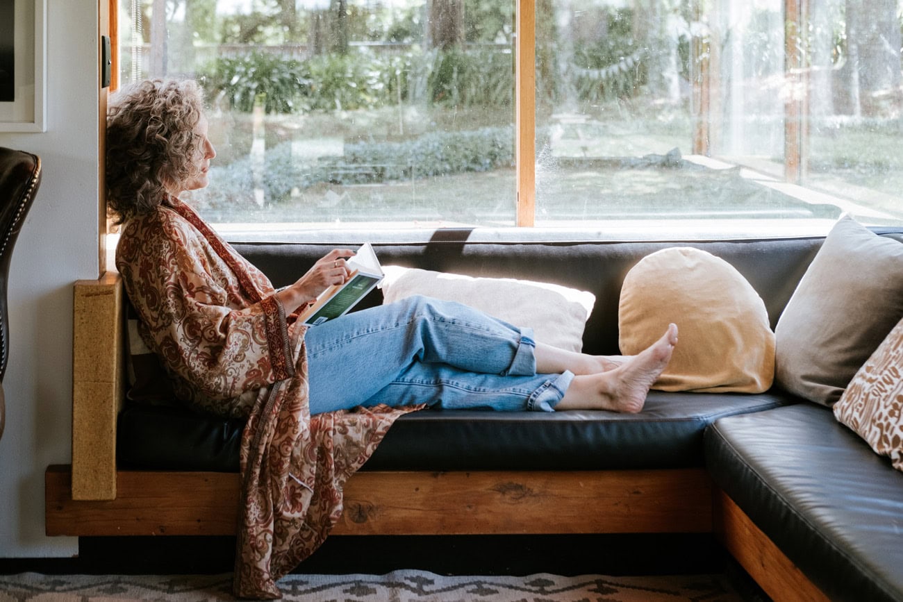 Photo by Florence Charvin Photography of A person with curly hair sits barefoot on a sofa by a window, reading a book. Cushions are placed along the sofa, and greenery is visible outside.