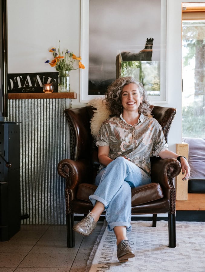 Photo by Florence Charvin Photography of A woman with curly hair sits in a leather armchair, smiling at the camera. Behind her is a shelf with flowers and a framed photo of a horse on the wall.