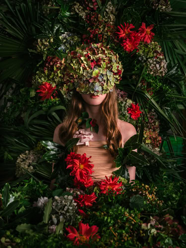 Photo by Florence Charvin Photography of A person stands surrounded by lush greenery and red flowers, their face mostly obscured by a large cluster of hydrangeas and other plants.