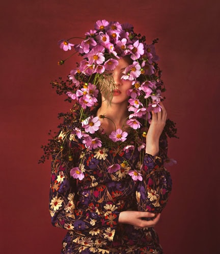 Photo by Florence Charvin Photography of A person in a floral dress stands against a burgundy background, with their face partially obscured by a cascade of pink flowers and green foliage.