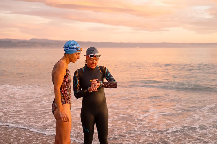 Photo by Florence Charvin Photography of Two swimmers in wetsuits and swim caps stand on a beach at sunset, with one checking a watch whilst the other looks on.