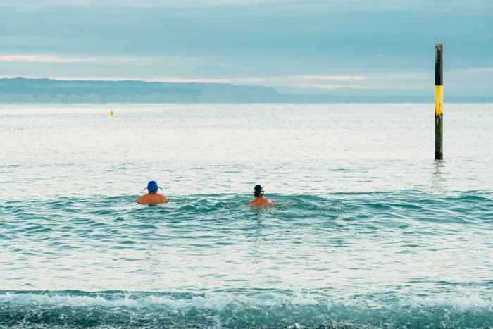 Photo by Florence Charvin Photography of Two people swim in the sea near a yellow and black pole, with distant cliffs and a cloudy sky in the background.
