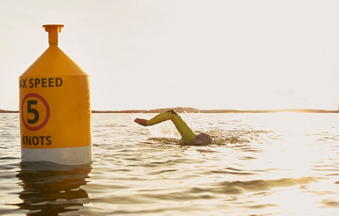 Photo by Florence Charvin Photography of A swimmer in a wetsuit passes a yellow buoy marked "MAX SPEED 5 KNOTS" on a body of water during daylight.