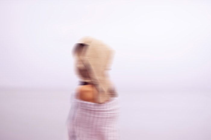 Photo by Florence Charvin Photography of A blurred image of a person wearing a hooded garment, facing sideways against a pale, indistinct background.