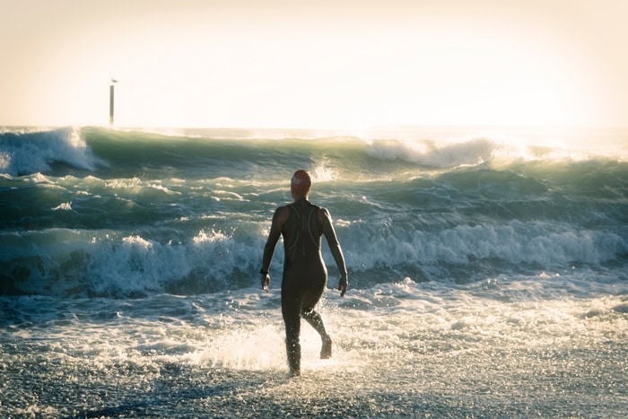 Photo by Florence Charvin Photography of Person in a wetsuit walks into the sea towards incoming waves under bright sunlight.
