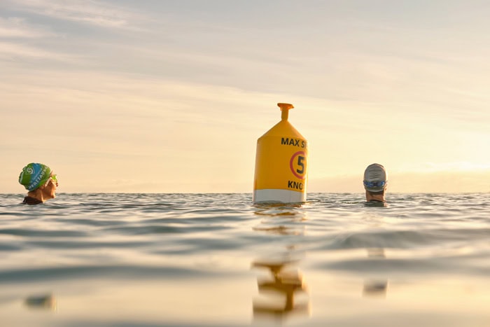 Photo by Florence Charvin Photography of Two swimmers in swimming caps are in calm water near a yellow buoy marked "MAX 5 KNOTS" under a clear sky.
