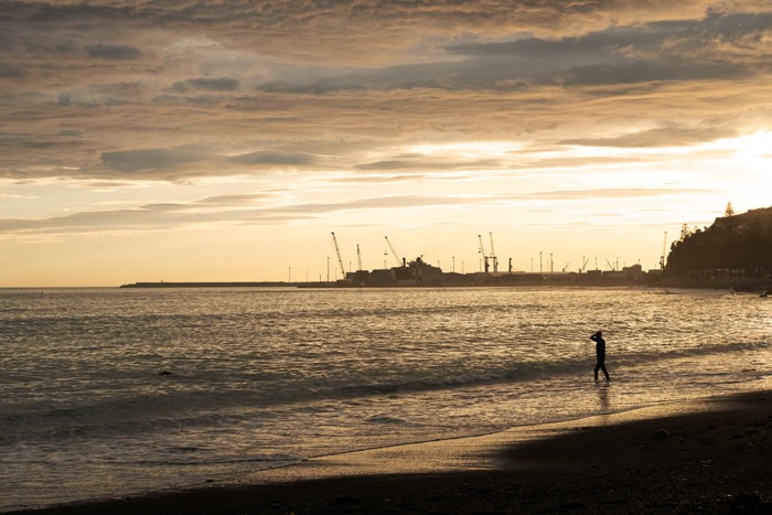 Photo by Florence Charvin Photography of A person stands at the edge of the water on a beach at sunset, with cranes and industrial structures visible in the background.